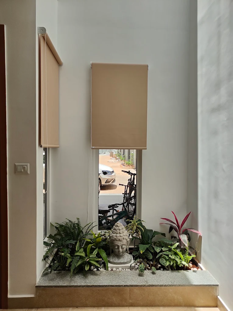 Beige roller blinds over a tall courtyard window, with plants and a stone Buddha below